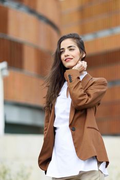 Young woman with nice hair standing outside of office building. Businesswoman wearing formal wear with wavy hairstyle. Young girl with brown jacket and trousers.