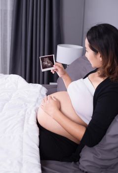 pregnant woman with ultrasound image of baby sitting on bed in the bedroom at home