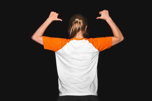 A girl in a T-shirt on a black background points with her hands to her back, a place for text. Design copy space clothes.