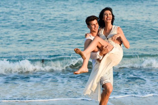 Young happy couple walking in a beautiful beach. Funny Man carrying a woman on his arms. People wearing casual clothes.