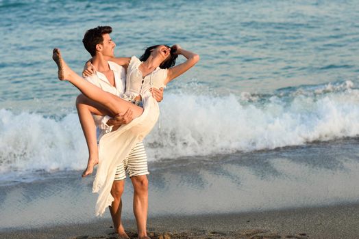 Young happy couple walking in a beautiful beach. Funny Man carrying a woman on his arms. People wearing casual clothes.