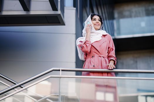 Young Muslim Woman walking on the street wearing hijab headscarf using smart phone in a office building. Business background.