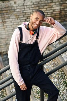 Young black man wearing casual clothes smiling on urban stairs. Happy African guy with bib pants outdoors