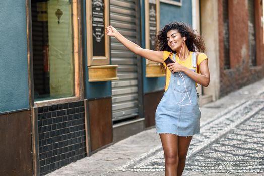 Attractive African woman listening to music with earphones outdoors. Black girl in casual clothes with curly hairstyle in urban background.