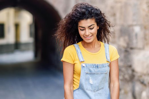 Young Arab woman with curly hairstyle outdoors. Arab girl in casual clothes in the street. Happy female wearing yellow t-shirt and denim dress in urban background.