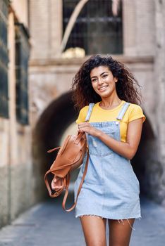 Young Arab tourist woman with curly hairstyle outdoors. Arab traveler girl in casual clothes in the street. Happy female wearing yellow t-shirt and denim dress in urban background.