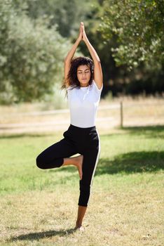 Young Arab woman doing yoga in nature. North African female wearing sport clothes doing tree figure in urban park.