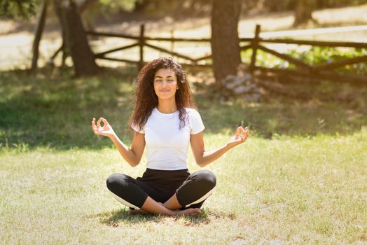 Young Arab woman doing yoga in nature. North African female wearing sport clothes doing lotus figure in urban park.