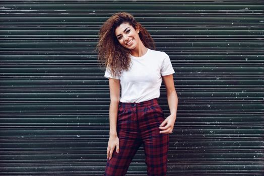 Smiling young arabic woman with black curly hairstyle. Arab girl in casual clothes in the street. Happy female wearing white t-shirt and checked pants against urban blinds.