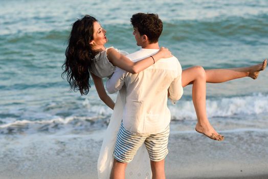 Young happy couple walking in a beautiful beach. Funny Man carrying a woman on his arms. People wearing casual clothes.