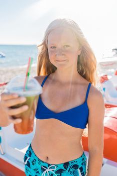 A young girl with blonde hair of European appearance, a teenager holds a colored cold non-alcoholic cocktail in her hand against the background of the sea beach.