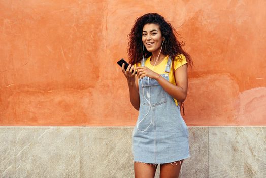 Happy Arab woman listening to music with earphones against red wall. African girl in casual clothes with curly hairstyle in urban background.