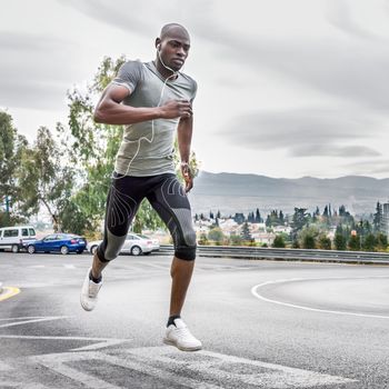 Black man running outdoors in urban road listening to music with white headphones. Young male exercising with city scape at the background.