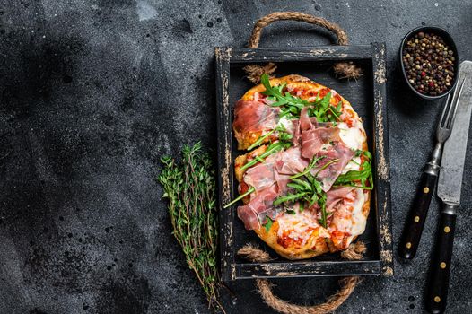 Italian Pizza with prosciutto parma ham, arugula salad and cheese in a rustic wooden tray. Black background. Top view. Copy space.