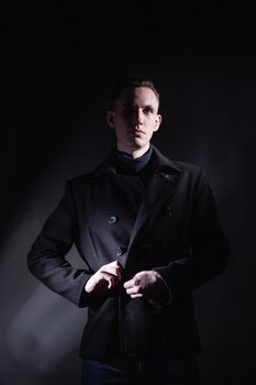 Serious confident young male in black coat looking away while standing under light in dark studio with black wall and shadow