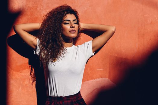 Beautiful young Arab woman with black curly hairstyle to the sun with her eyes closed. Female in casual clothes in the street.