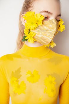 Slim female model in protective mask with fresh yellow flowers standing in transparent outfit on white background in studio