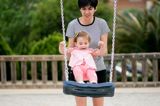 Mother swinging her little daughter on a swing in a playground