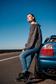 Young woman near roofless car and smiling outdoors on sunny day near the empty road. No people, sportive blue car