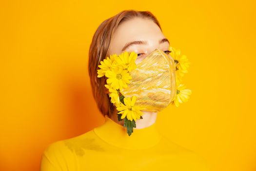 Slim female model in protective mask with fresh yellow flowers standing in transparent outfit on yellow background in studio