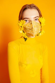 Slim female model in protective mask with fresh yellow flowers standing in transparent outfit on yellow background in studio