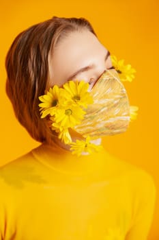 Slim female model in protective mask with fresh yellow flowers standing in transparent outfit on yellow background in studio