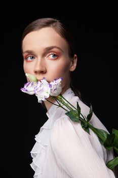 Pretty young woman in elegant white blouse touching face with natural flowers and looking away against black background