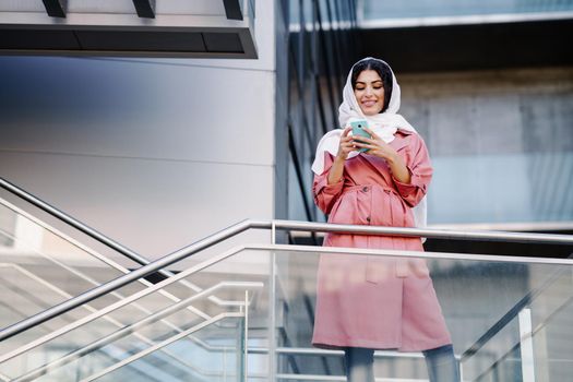 Young Arab woman wearing hijab headscarf texting message with her smartphone in a office building. Business background.