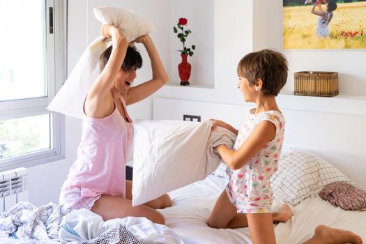 Middle-age mother with her eight years daughter having funny pillow fight on bed. Mum spending free time with her daughter.