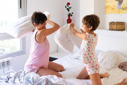 Middle-age mother with her eight years daughter having funny pillow fight on bed. Mum spending free time with her daughter.