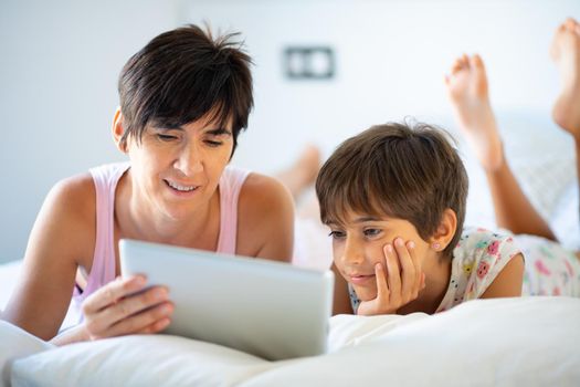 Middle-age mother with her eight years daughter using digital tablet in bedroom. Funny mom and lovely little girl are having fun on the bed.