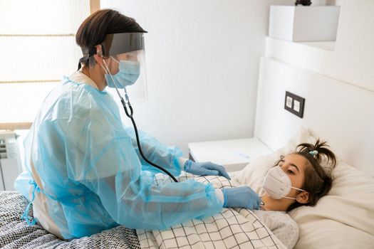 Doctor protected by personal protective equipment examining a little girl with a stethoscope at home.