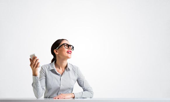 Smiling young woman sits at desk and dreamy looking upward. Portrait of beautiful girl in glasses with bright red lips on white wall background. Human resource and career development concept.