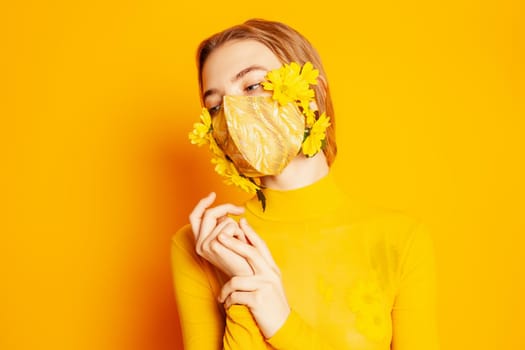 Slim female model in protective mask with fresh yellow flowers standing in transparent outfit on yellow background in studio