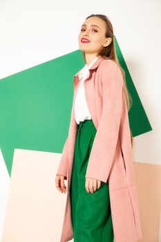 Confident young female model in elegant pastel pink coat and bright green pants representing spring fashion looking away in studio