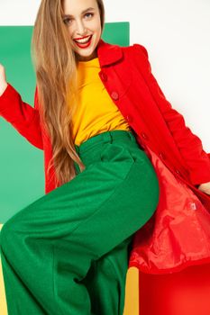 Coquettish young female model in elegant vivid multicolored green red yellow clothes standing against colorful background in studio