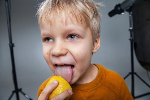Crop cute little boy with blond hair licking fresh ripe sour lemon in gray studio