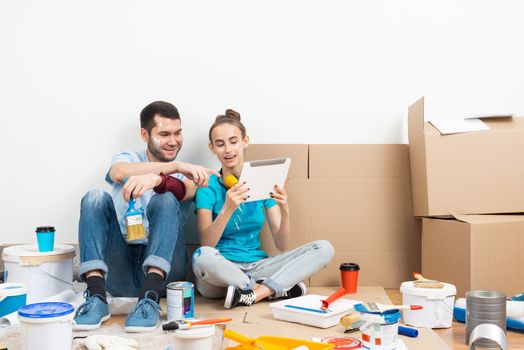 Happy couple watching something funny on tablet computer while sitting on floor. Painting tools and materials for home remodeling and interior redesign. Young family renovating their home after moving