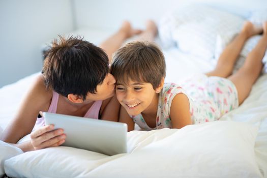 Middle-age mother with her eight years daughter using digital tablet in bedroom. Funny mom and lovely little girl are having fun on the bed.