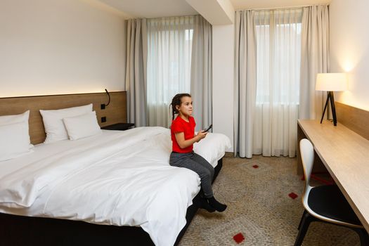 Little girl child with remote control watching tv set lying on bed in room at home alone. cute little girl