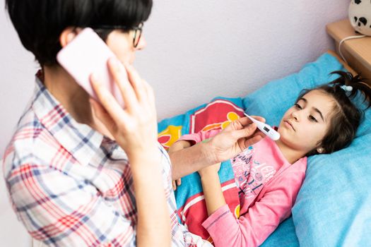 Mother worried about her daughter's temperature calling the doctor. Little girl on bed.