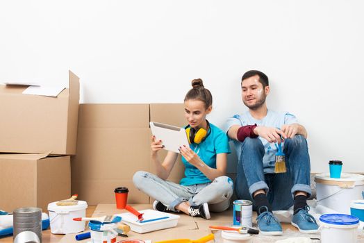 Happy couple sitting on floor and looking at tablet computer screen. Home remodeling and house interior redesign. Painting tools and materials on floor. Young family renovating their home after moving