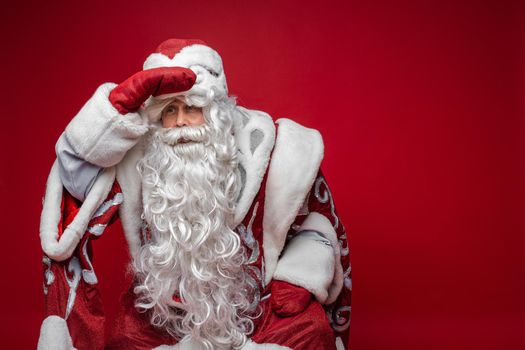 santa claus with long white beard tries to see someone, picture isolated on red background