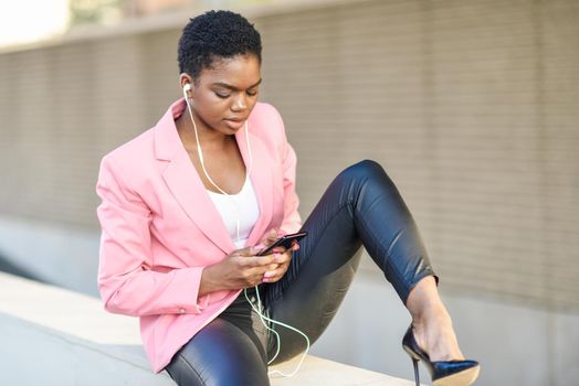 Black businesswoman sitting outdoors using smartphone with earphones. African american female wearing suit with pink jacket.