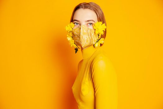 Slim female model in protective mask with fresh yellow flowers standing in transparent outfit on yellow background in studio