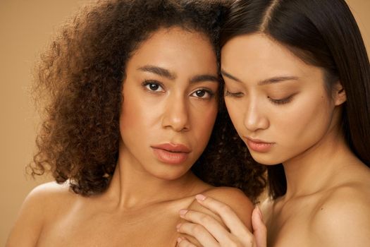 Face closeup of two sensual beautiful mixed race young women with perfect skin posing for camera isolated over beige background. Skincare, diversity concept. Selective focus