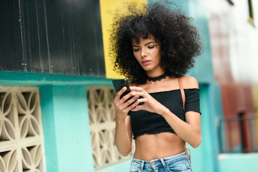 Young black woman with afro hair walking on the street holding a smart phone. Female wearing casual clothes in urban background. Lifestyle concept