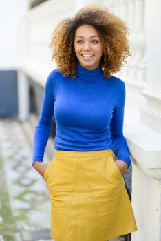 Beautiful young African American woman, model of fashion, smiling with afro hairstyle and green eyes wearing blue sweater and yellow skirt in urban background