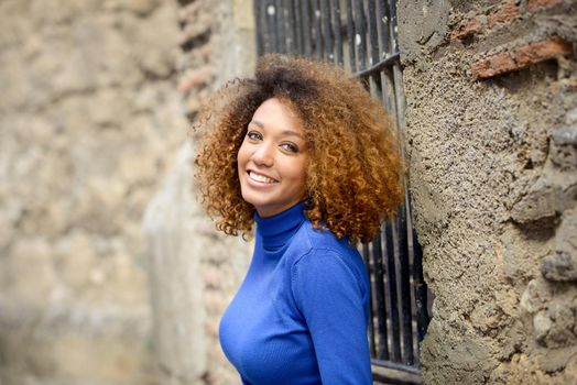 Beautiful young African American woman, model of fashion, smiling with afro hairstyle and green eyes wearing blue sweater in urban background