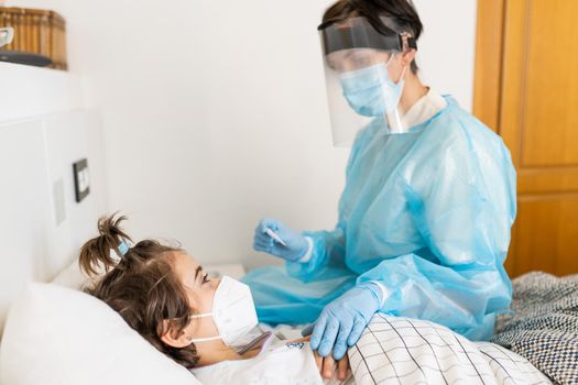 Doctor protected by personal protective equipment examining a little girl with a stethoscope at home.